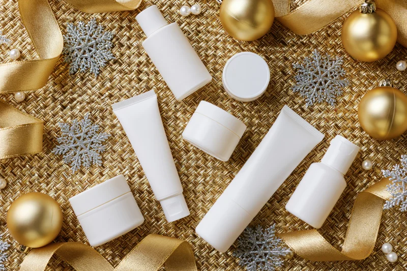 White skincare product tubes jars and bottles arranged on a golden woven surface with holiday ornaments and ribbons