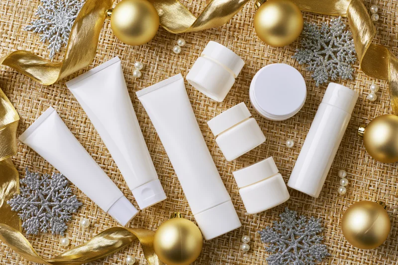 White skincare product tubes jars and bottles arranged with gold ornaments and silver snowflakes on textured burlap background