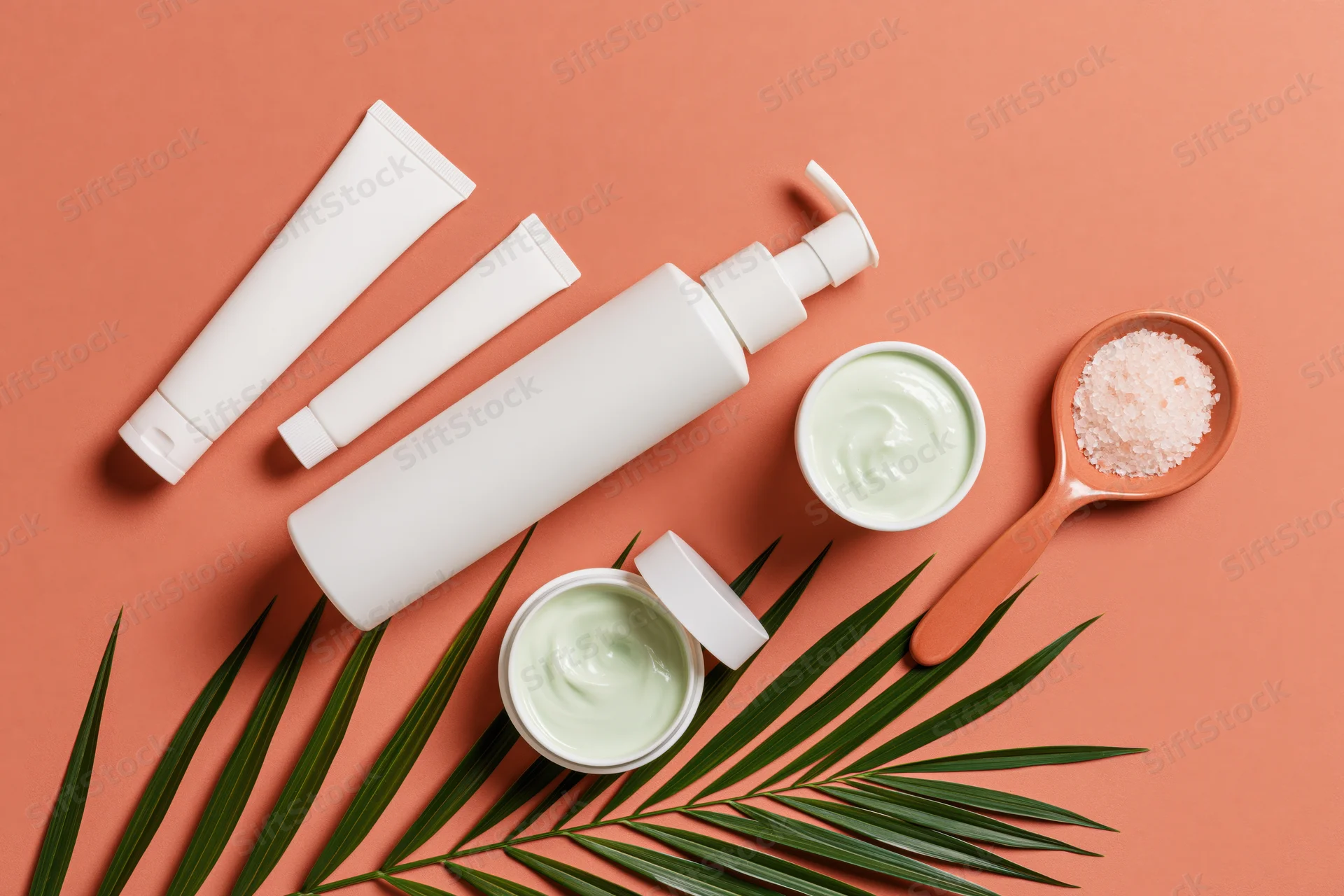White unlabeled skincare tubes and jars with palm leaf on coral background