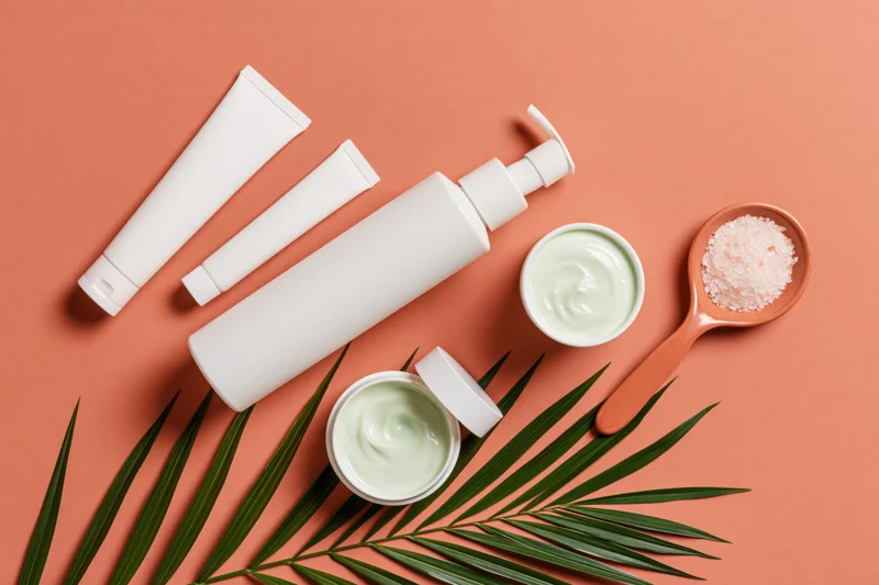 White unlabeled skincare tubes and jars with palm leaf on coral background