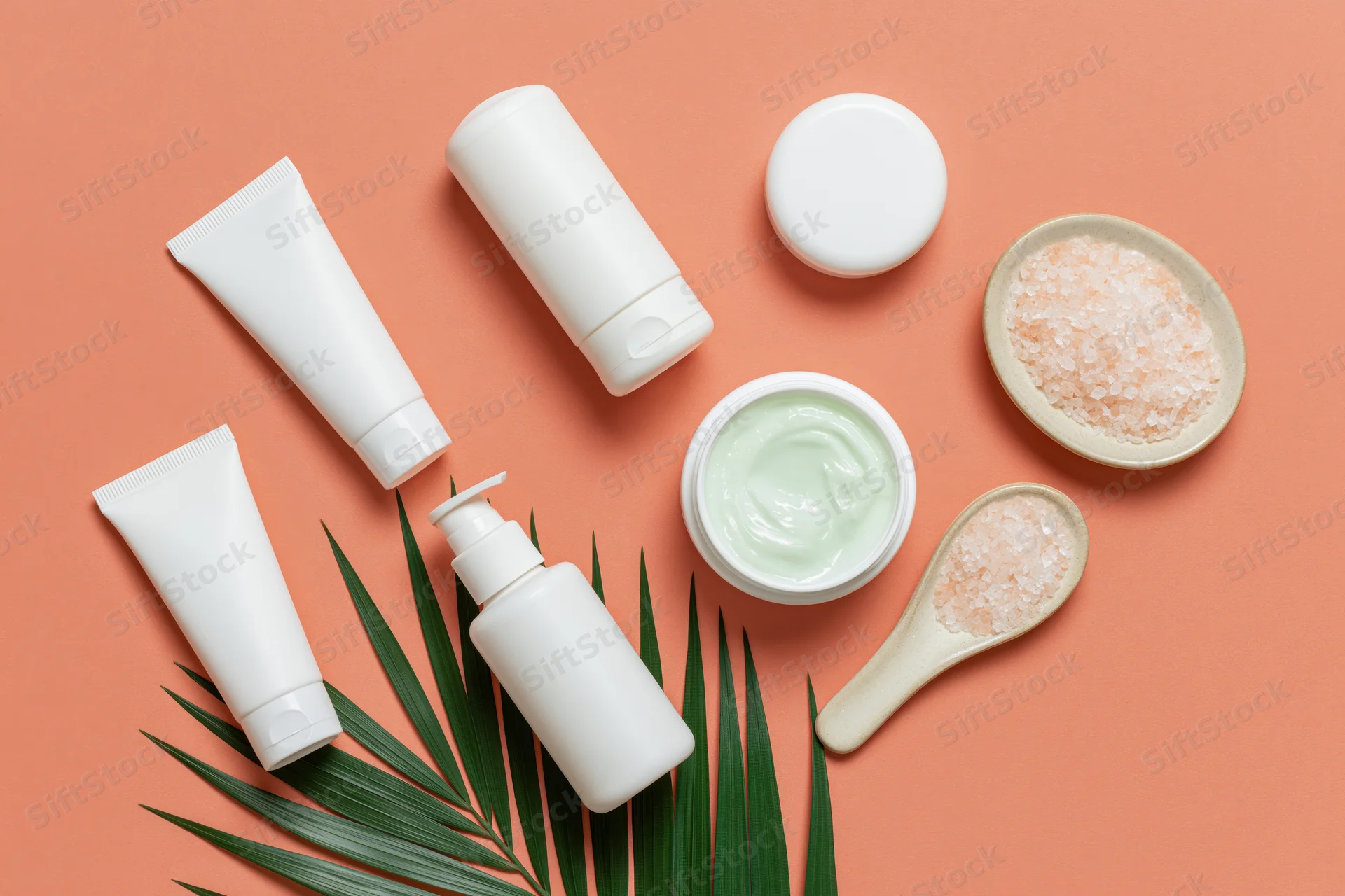 White unlabeled skincare tubes and jars with salt and palm leaf on orange background