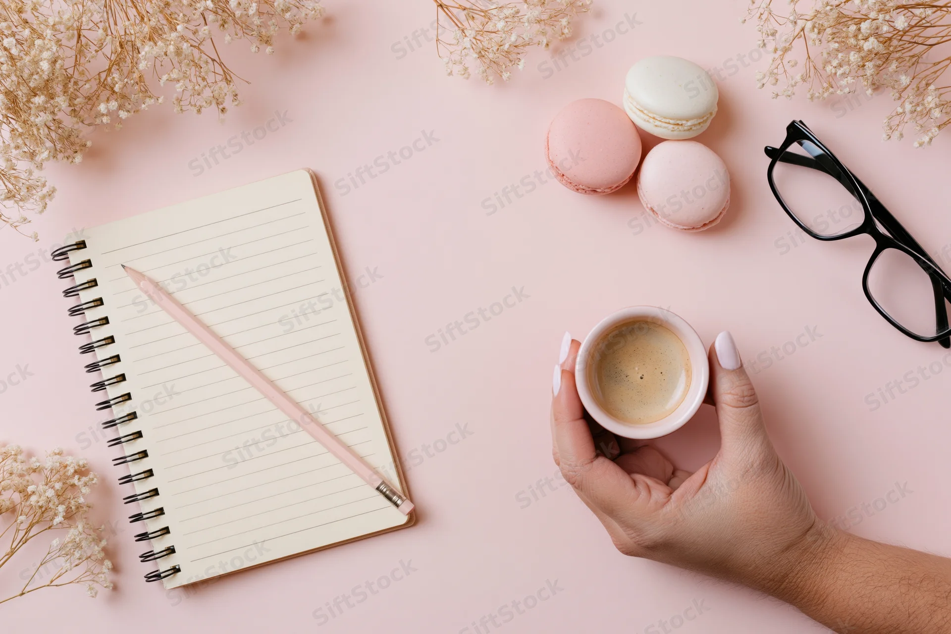 Hand holding coffee cup on pastel desk with notebook, pencil, macarons, glasses and dried flowers