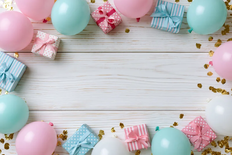 Pastel balloons and wrapped gifts arranged around a white wooden table with gold confetti, forming a border with central copyspace