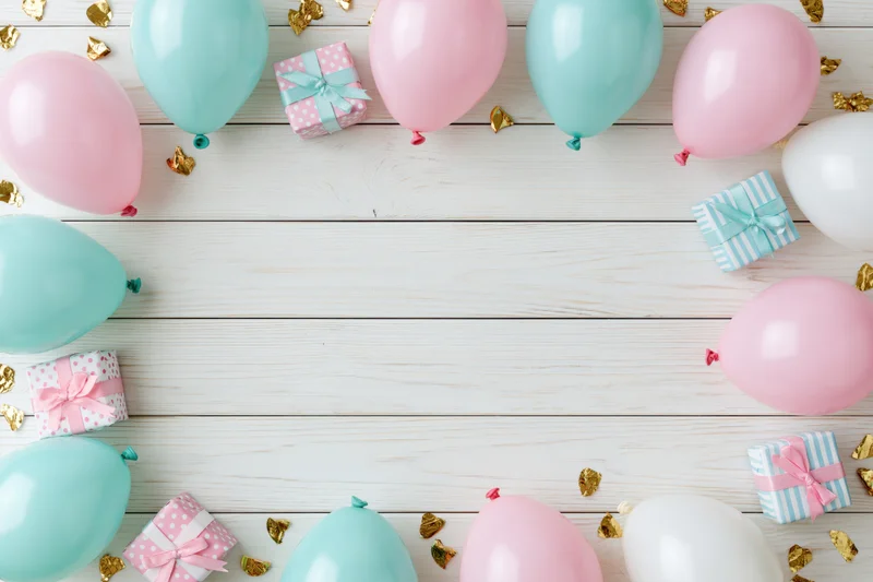 Pastel balloons and wrapped gifts arranged around a white wooden surface with gold confetti for a birthday party flat lay