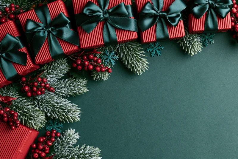 Red striped gift boxes with dark green ribbons and pine branches arranged along the top and left edge on a dark green surface