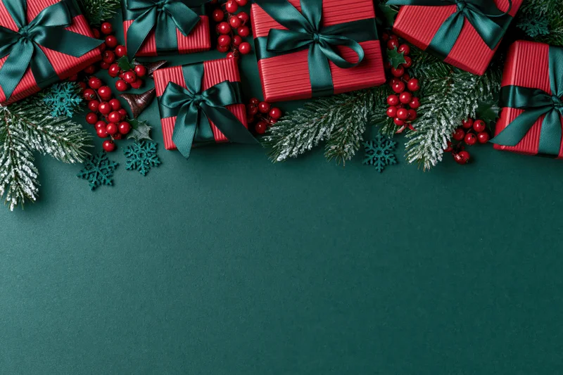 Red striped gift boxes with dark green ribbons and pine branches arranged along the top of a dark green surface, leaving copyspace below