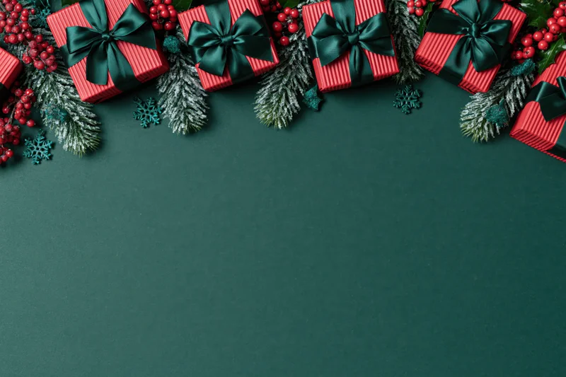 Red striped gift boxes with dark green bows and pine branches arranged along top edge on dark green surface with copyspace below