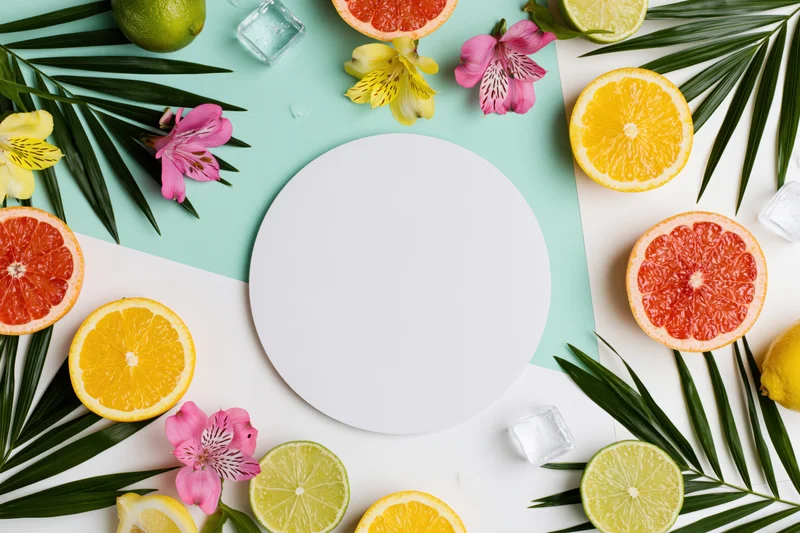 Flat lay arrangement of citrus fruits, tropical leaves and flowers around a blank white circular card on a colorful background
