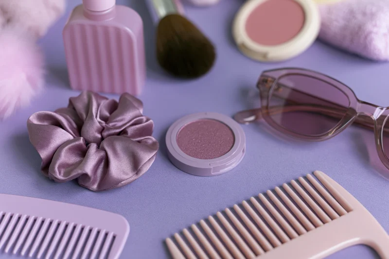 Beauty accessories arranged in flatlay on purple surface with combs, scrunchie, sunglasses and makeup products