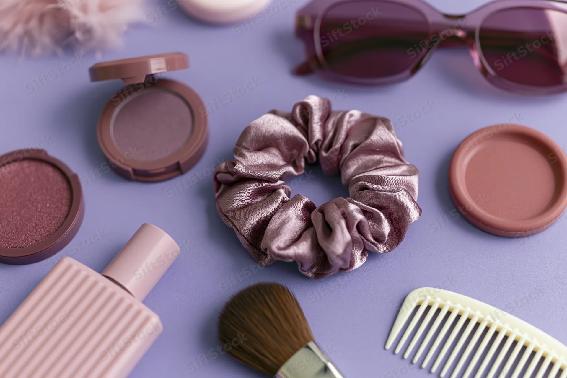 Beauty accessories arranged in flatlay on purple surface including makeup, hair scrunchie, brush, comb and sunglasses