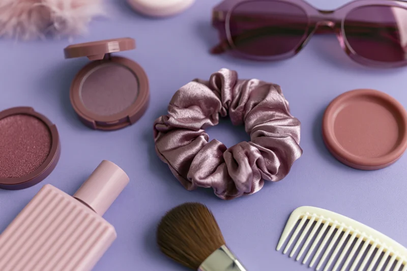 Beauty accessories arranged in flatlay on purple surface including makeup, hair scrunchie, brush, comb and sunglasses