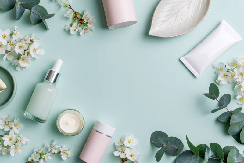 Skincare products arranged in a border frame with flowers and leaves on a mint background