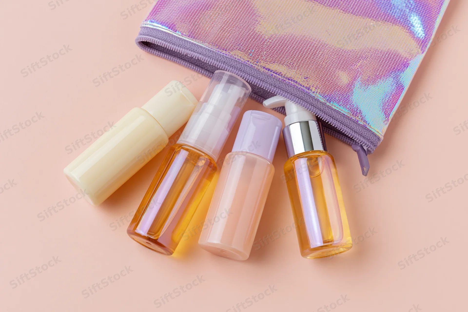 Travel-size skincare bottles and a pump dispenser spilling from an iridescent pouch on a pastel surface