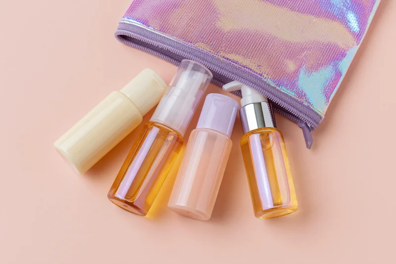 Travel-size skincare bottles and a pump dispenser spilling from an iridescent pouch on a pastel surface