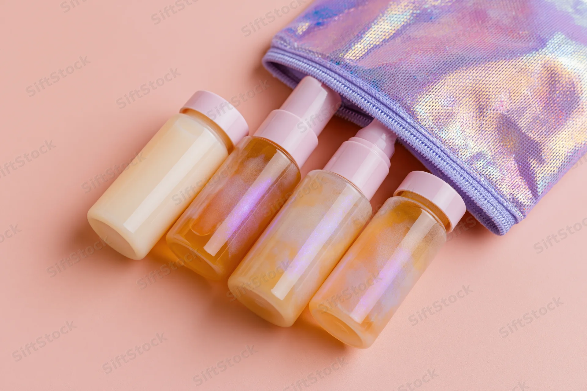 Four travel-size skincare bottles and a pump dispenser spilling from an iridescent pouch on a pastel pink surface