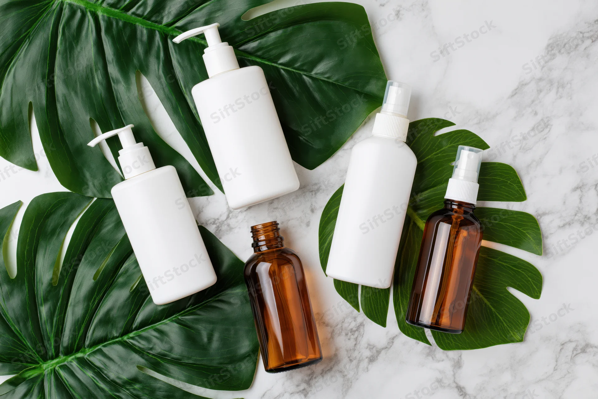 Skincare bottles with pump and spray dispensers arranged on monstera leaves and marble surface