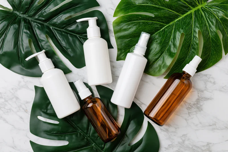 Skincare bottles with pump and spray dispensers arranged on monstera leaves and marble surface