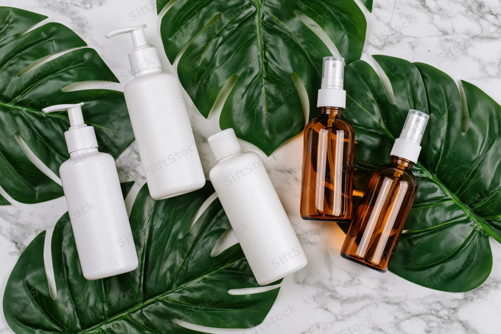 Skincare bottles with pump and spray dispensers arranged on monstera leaves and marble surface