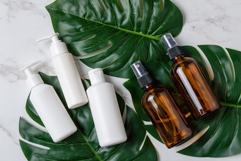 Skincare bottles with pump and spray dispensers arranged on monstera leaves on marble surface