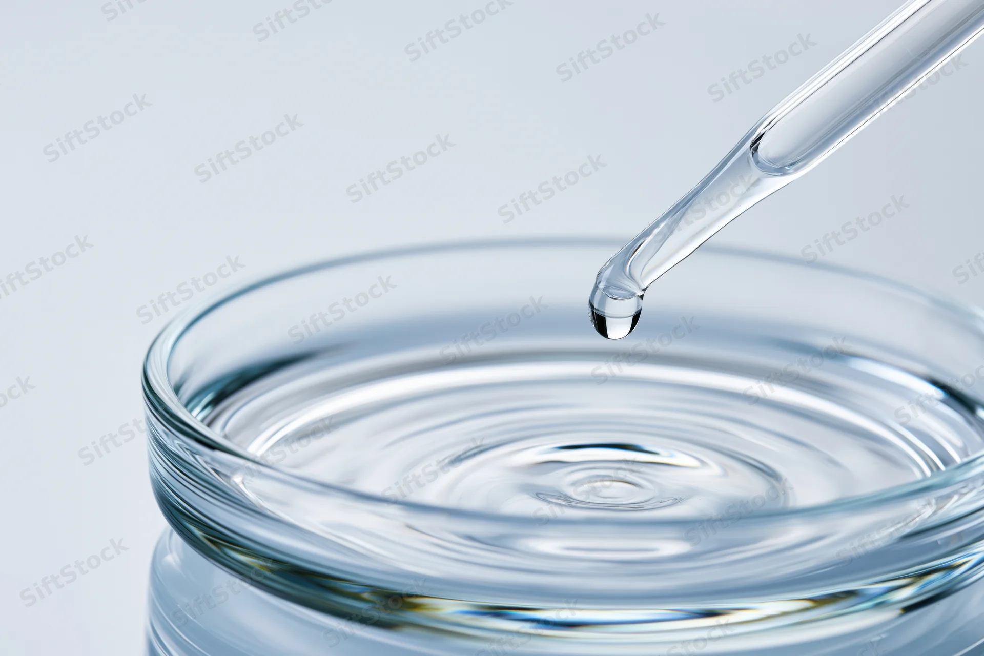 Glass pipette releasing a drop of clear liquid onto a round glass dish, creating ripples on the surface