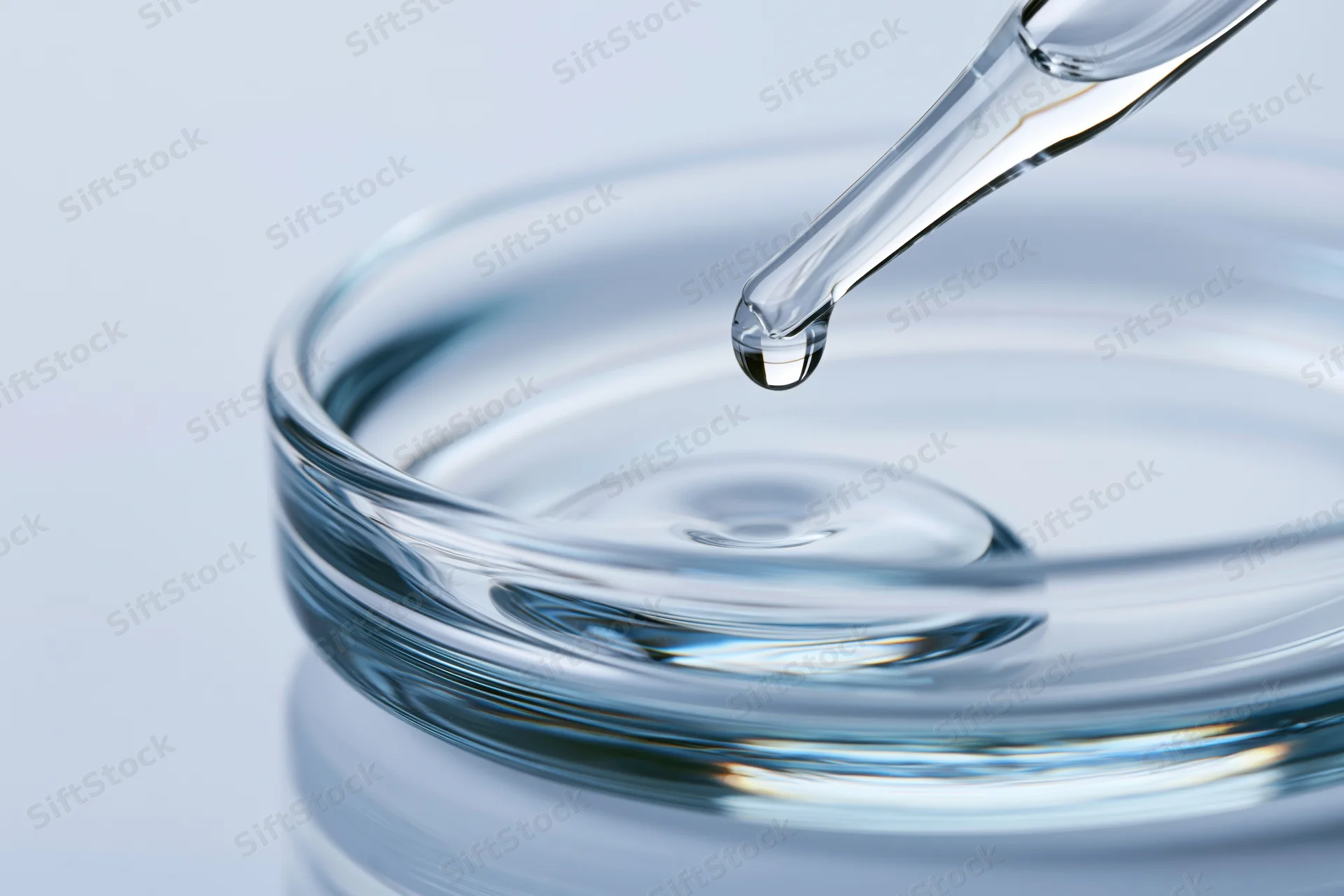 Glass pipette releasing a drop of clear liquid into a shallow glass dish
