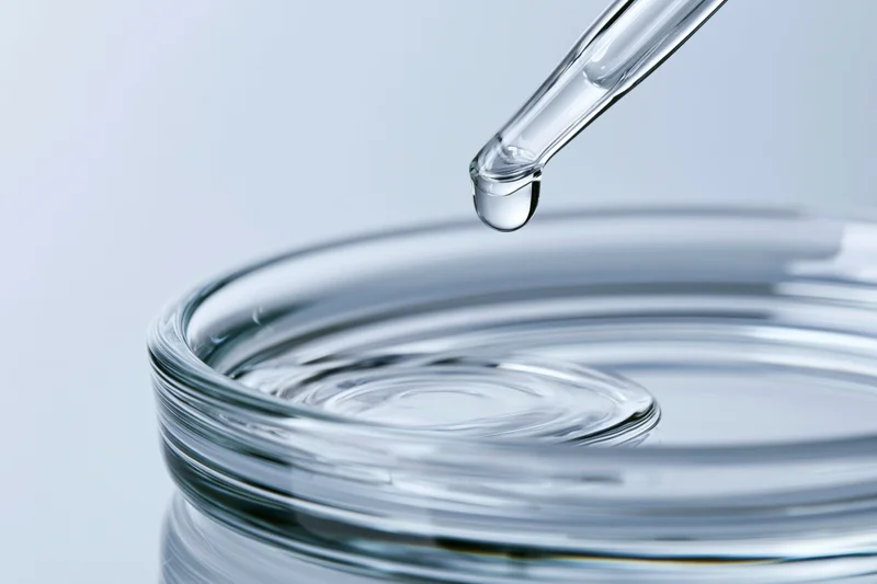 Glass pipette releasing a drop of clear liquid onto a round glass container surface