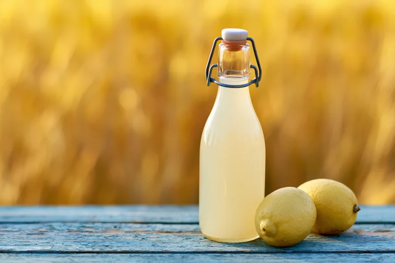 Glass bottle of pale lemon juice with lemons on a blue wooden table outdoors