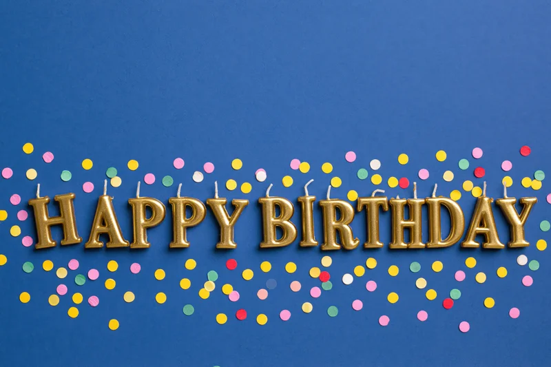 Gold letter-shaped birthday candles spelling happy birthday on blue background with colorful confetti