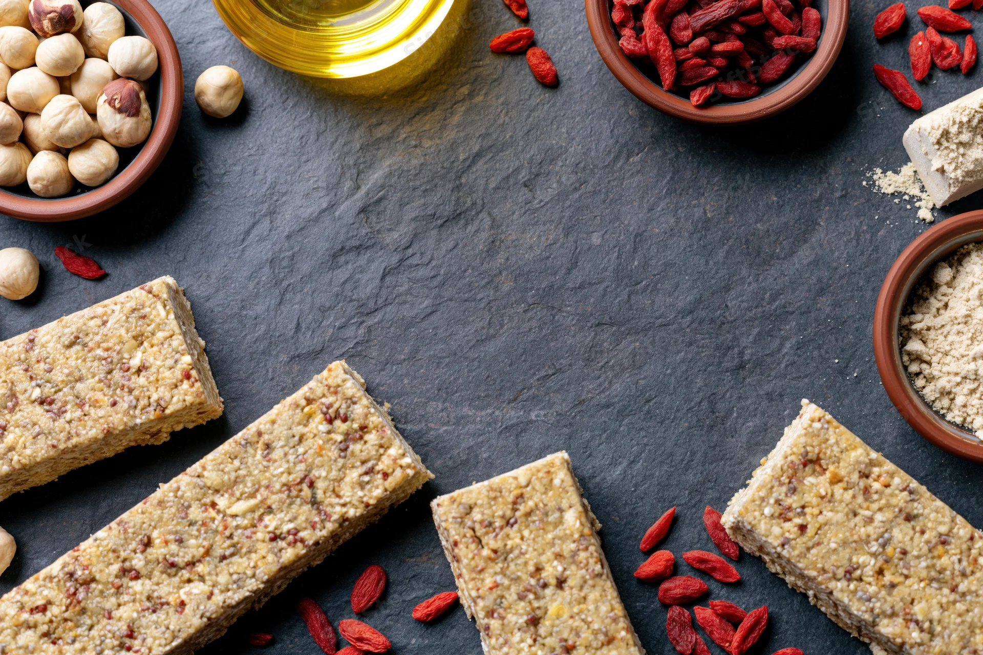 Granola bars arranged in a loose frame around scattered goji berries, hazelnuts, oil, and powder on dark slate surface