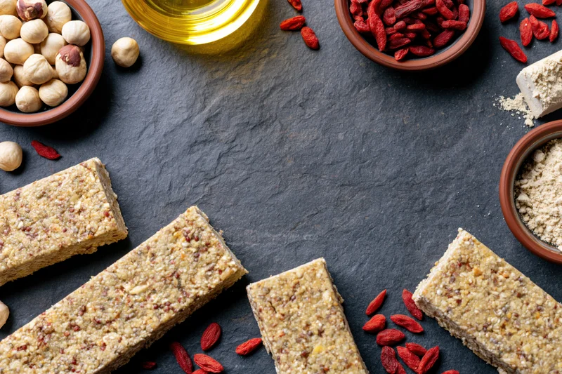 Granola bars arranged in a loose frame around scattered goji berries, hazelnuts, oil, and powder on dark slate surface