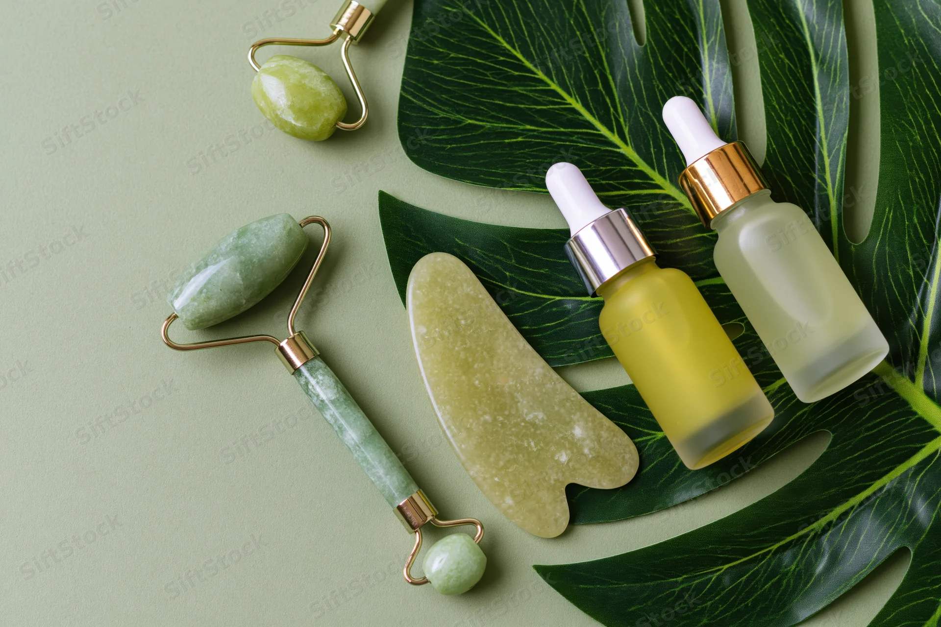 Jade facial rollers and serum bottles arranged with monstera leaves on a green surface