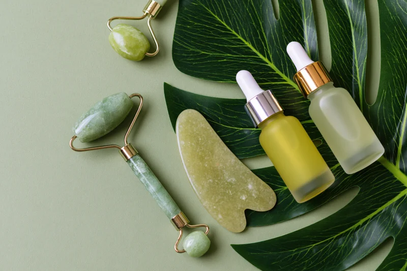 Jade facial rollers and serum bottles arranged with monstera leaves on a green surface