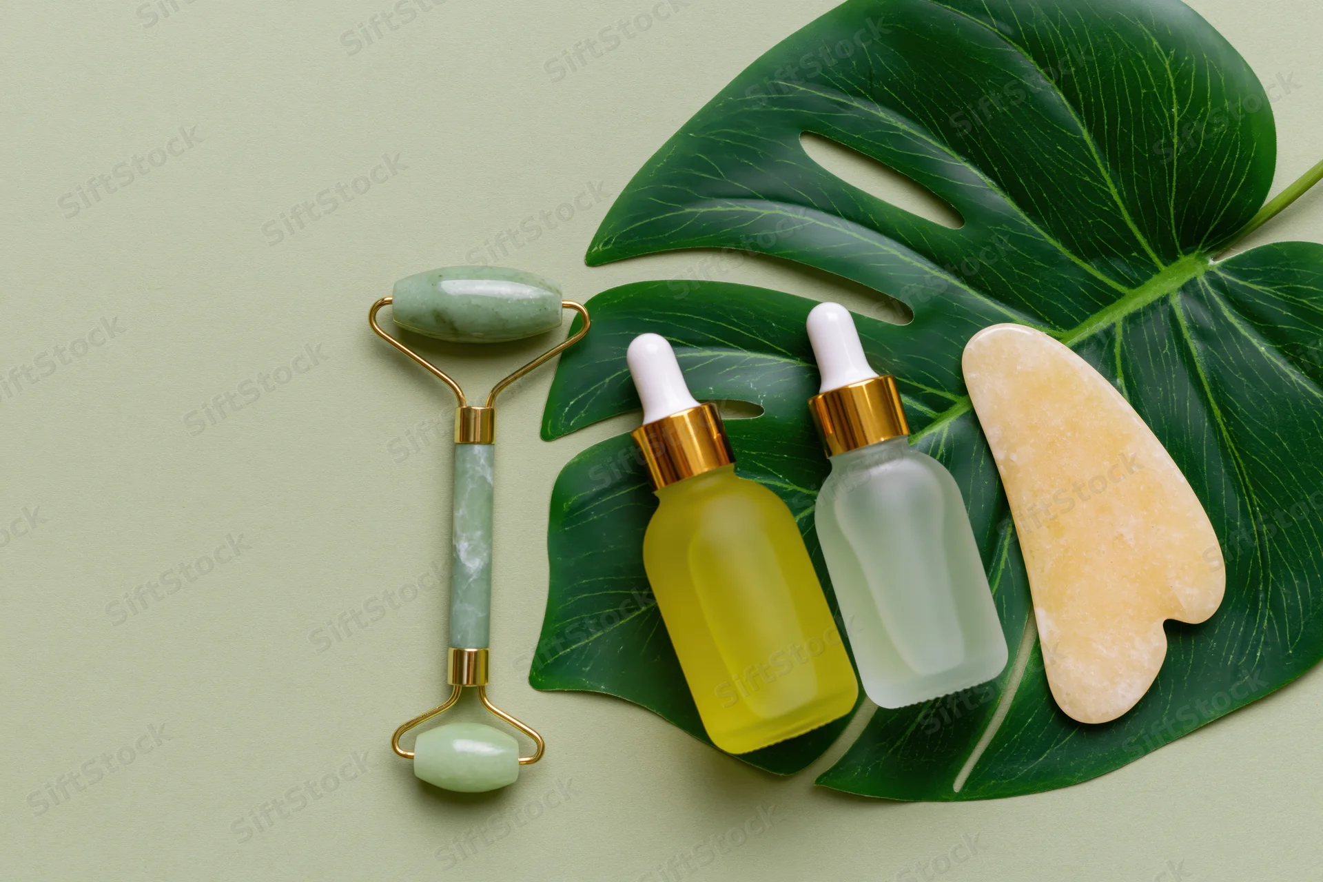Jade facial roller and two dropper serum bottles arranged on a monstera leaf with copyspace on a pale green background
