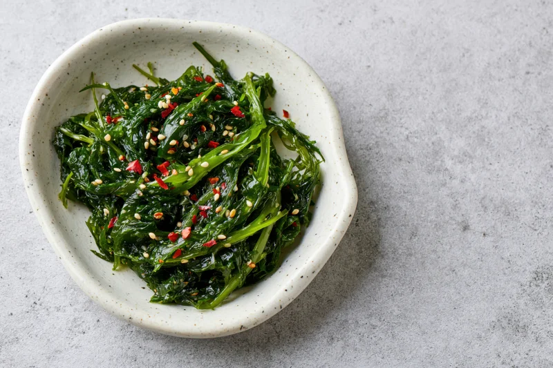 Wakame seaweed salad with sesame seeds and chili flakes in a white ceramic bowl on a gray textured surface