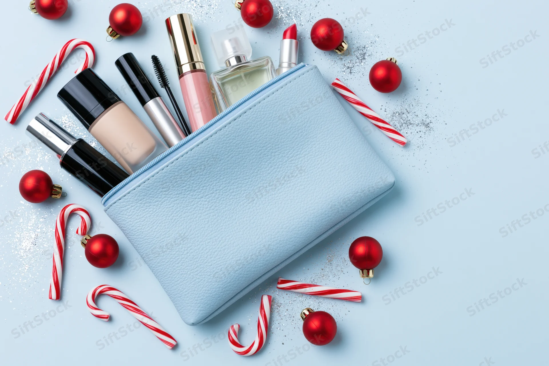 Open light blue cosmetics pouch with makeup products and red holiday ornaments on a light blue surface