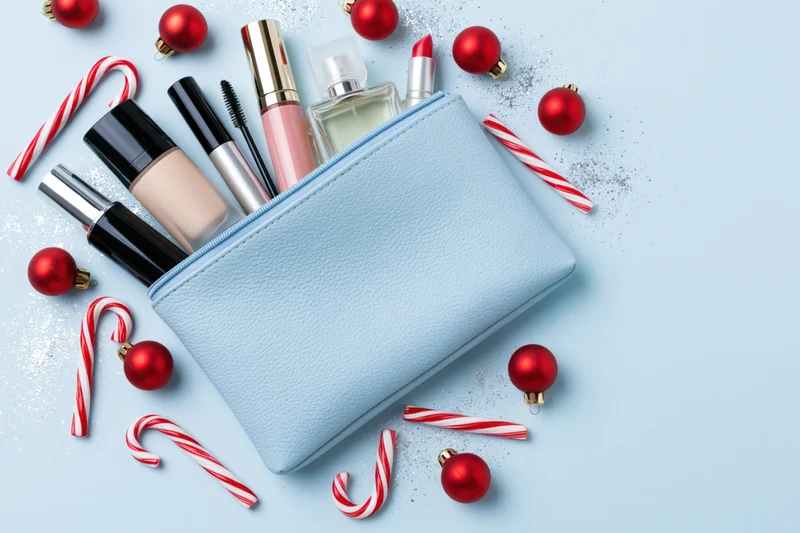 Open light blue cosmetics pouch with makeup products and red holiday ornaments on a light blue surface