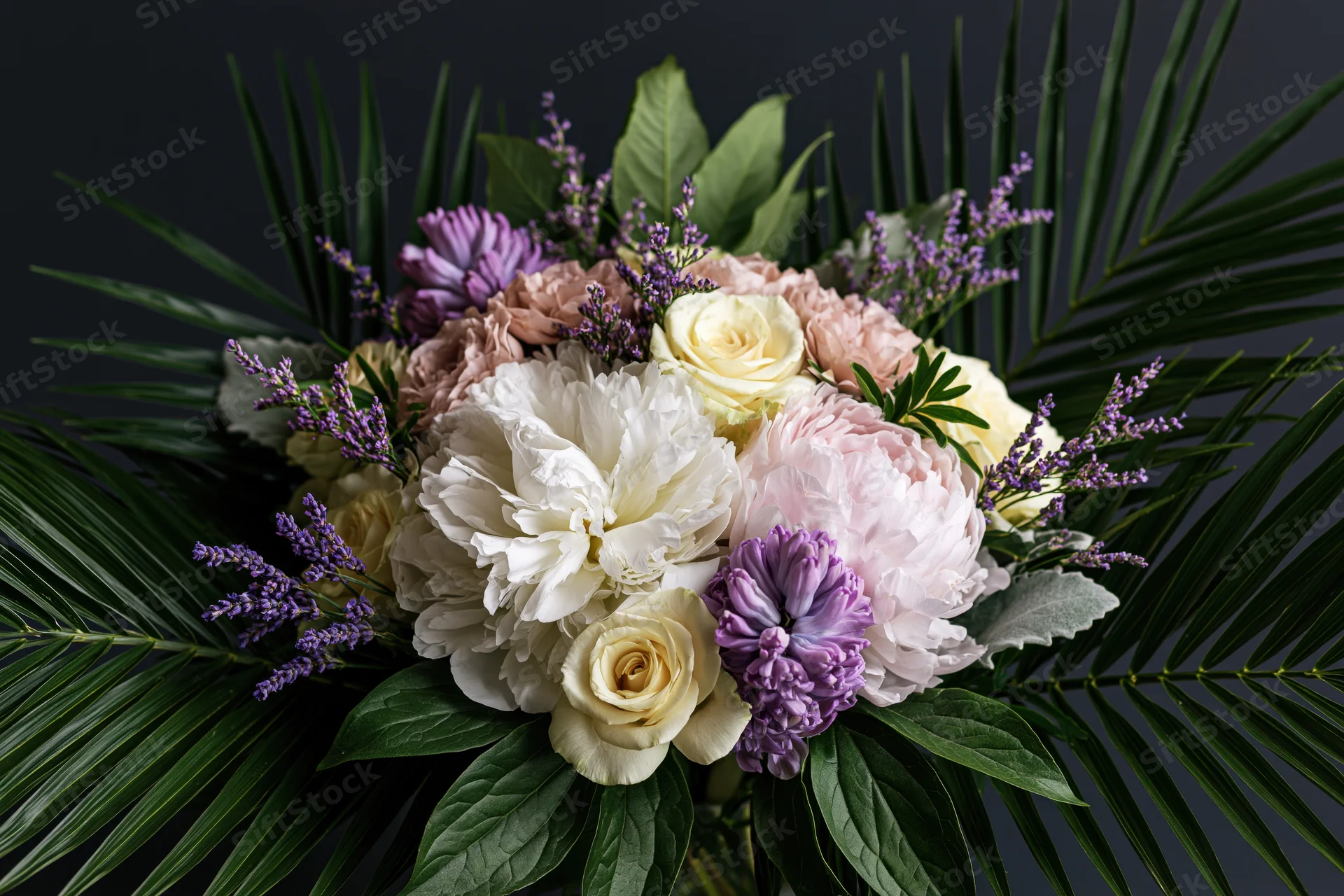 Lush floral arrangement with white and pink peonies, yellow roses, purple lavender, and green palm fronds against dark background