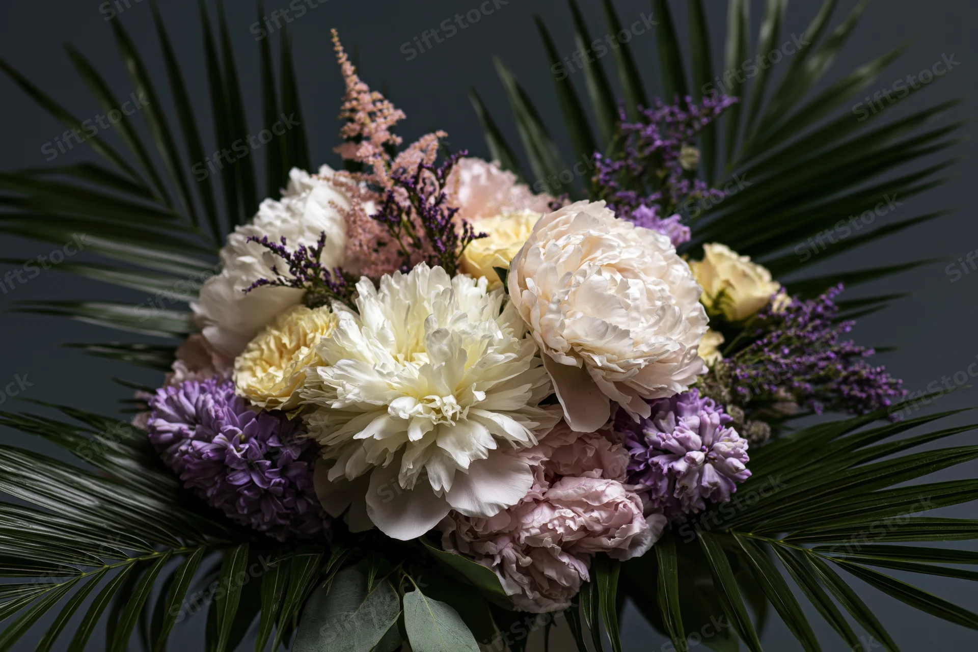 Lush floral arrangement with white and blush peonies, purple dahlias, and green palm leaves against dark background