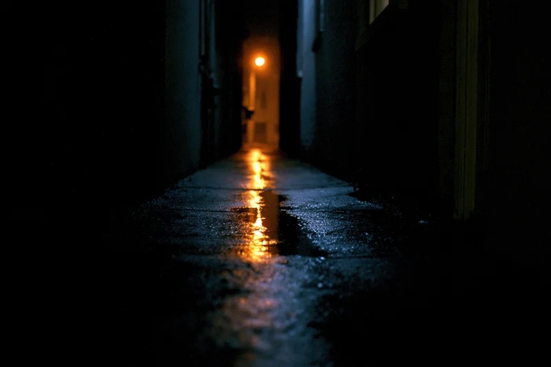 Narrow urban alley at night with wet concrete reflecting distant orange light