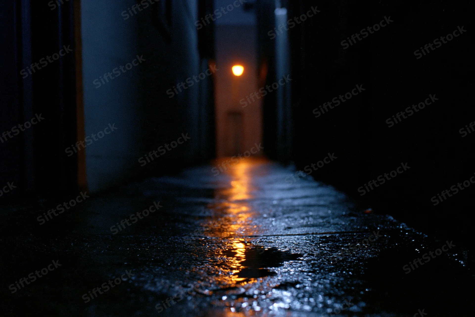 Narrow urban alley at night with wet concrete pavement reflecting distant orange light