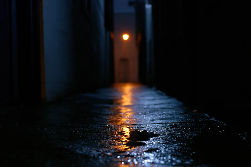 Narrow urban alley at night with wet concrete pavement reflecting distant orange light