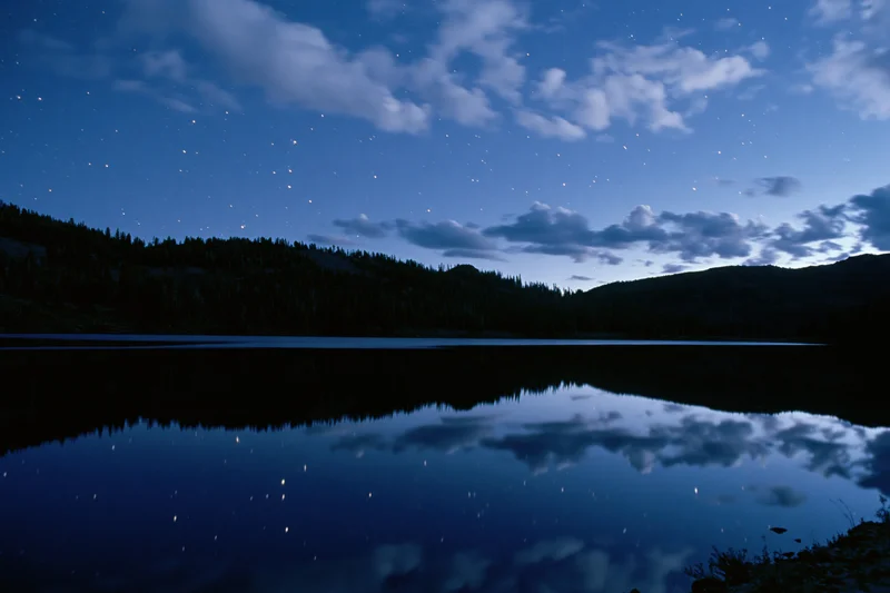 Night sky with stars reflected in calm mountain lake surrounded by forested hills