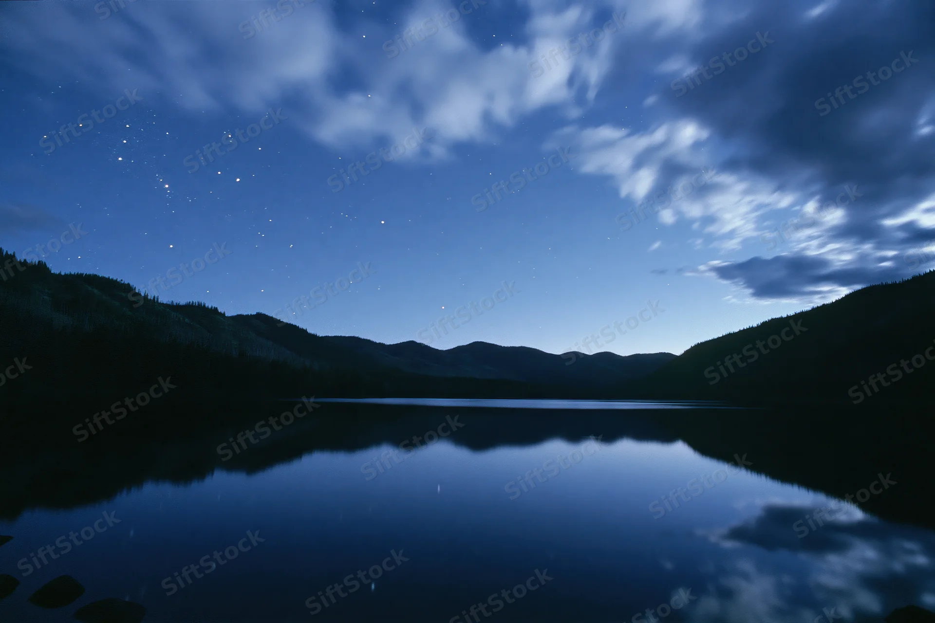 Starry night sky reflected in calm mountain lake surrounded by forested hills