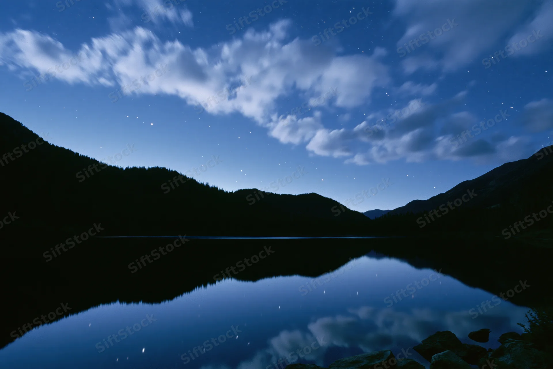 Starry night sky reflected in calm mountain lake surrounded by dark forested hills