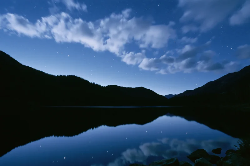 Starry night sky reflected in calm mountain lake surrounded by dark forested hills
