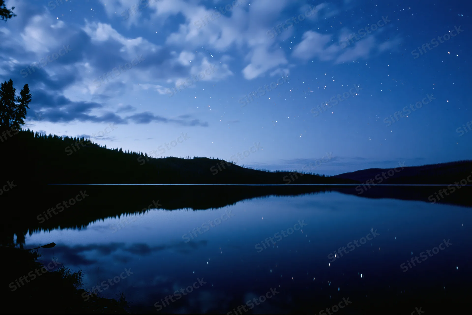 Night sky with stars reflected in calm mountain lake surrounded by forested hills
