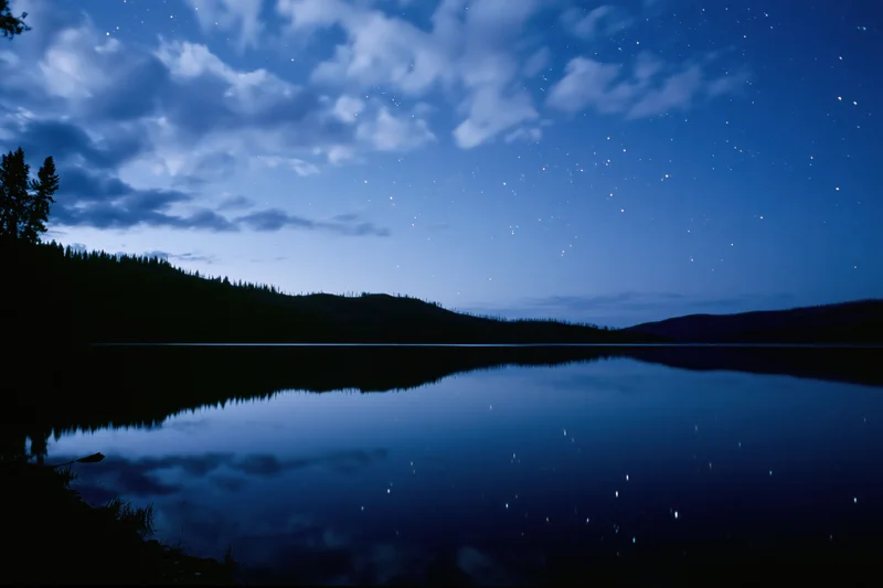 Night sky with stars reflected in calm mountain lake surrounded by forested hills