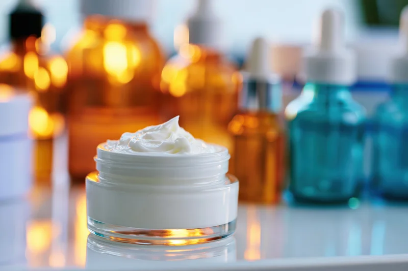 Open glass jar of white face cream centered on reflective surface with skincare bottles in background