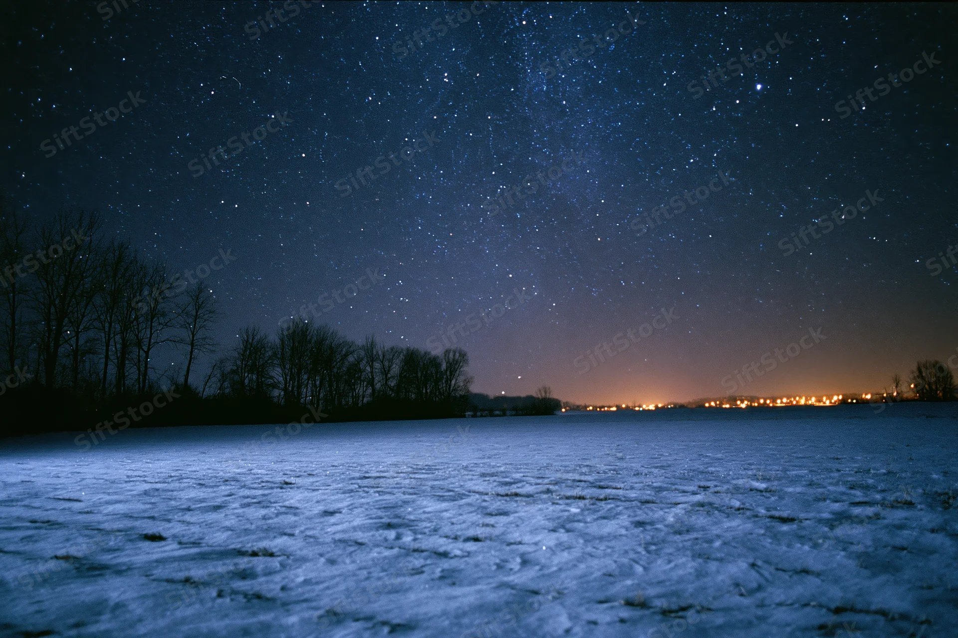 Snow-covered field under star-filled night sky with distant town lights on horizon