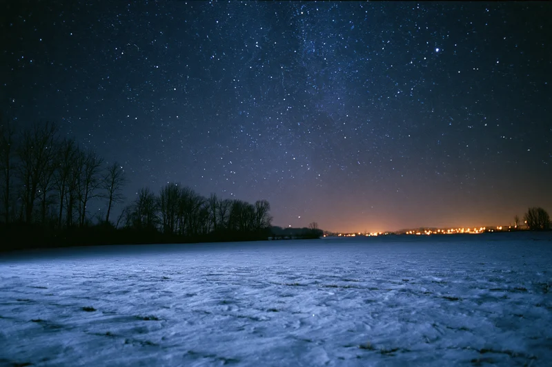 Snow-covered field under star-filled night sky with distant town lights on horizon